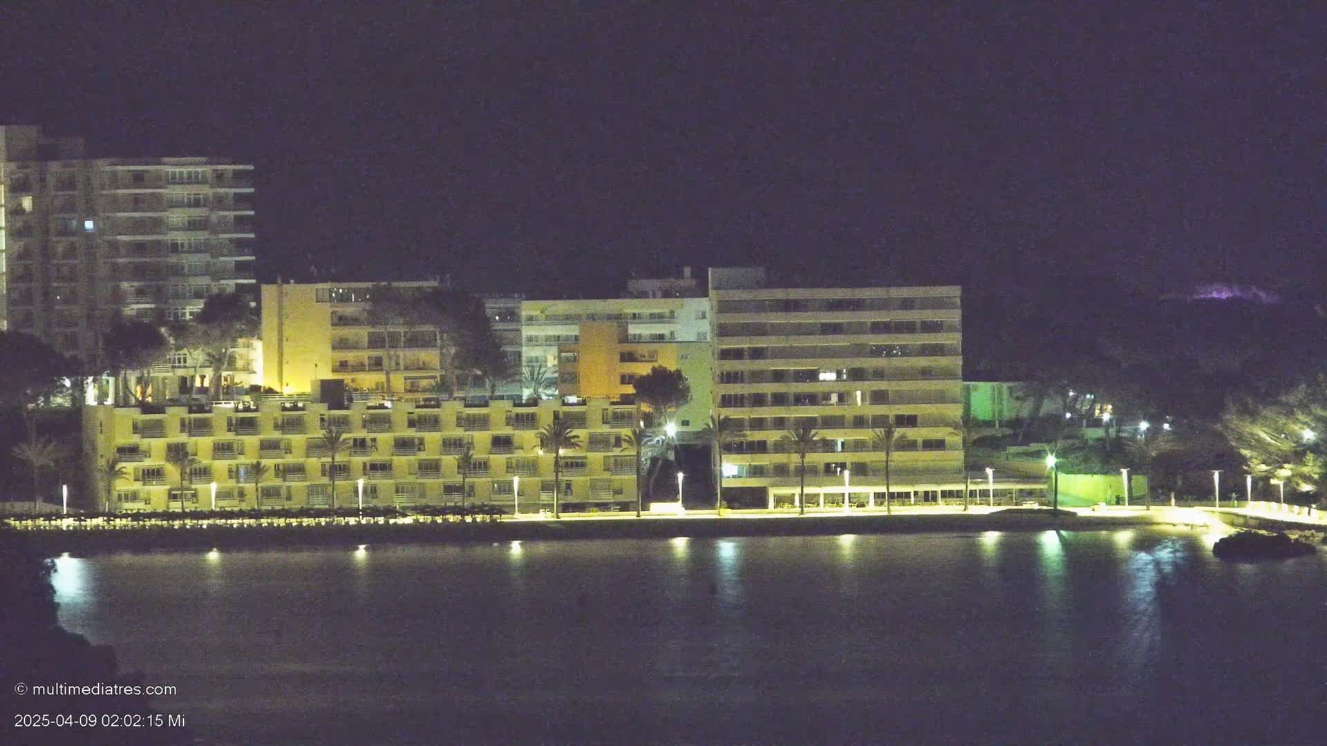 A row of illuminated buildings stands along a calm body of water at night.