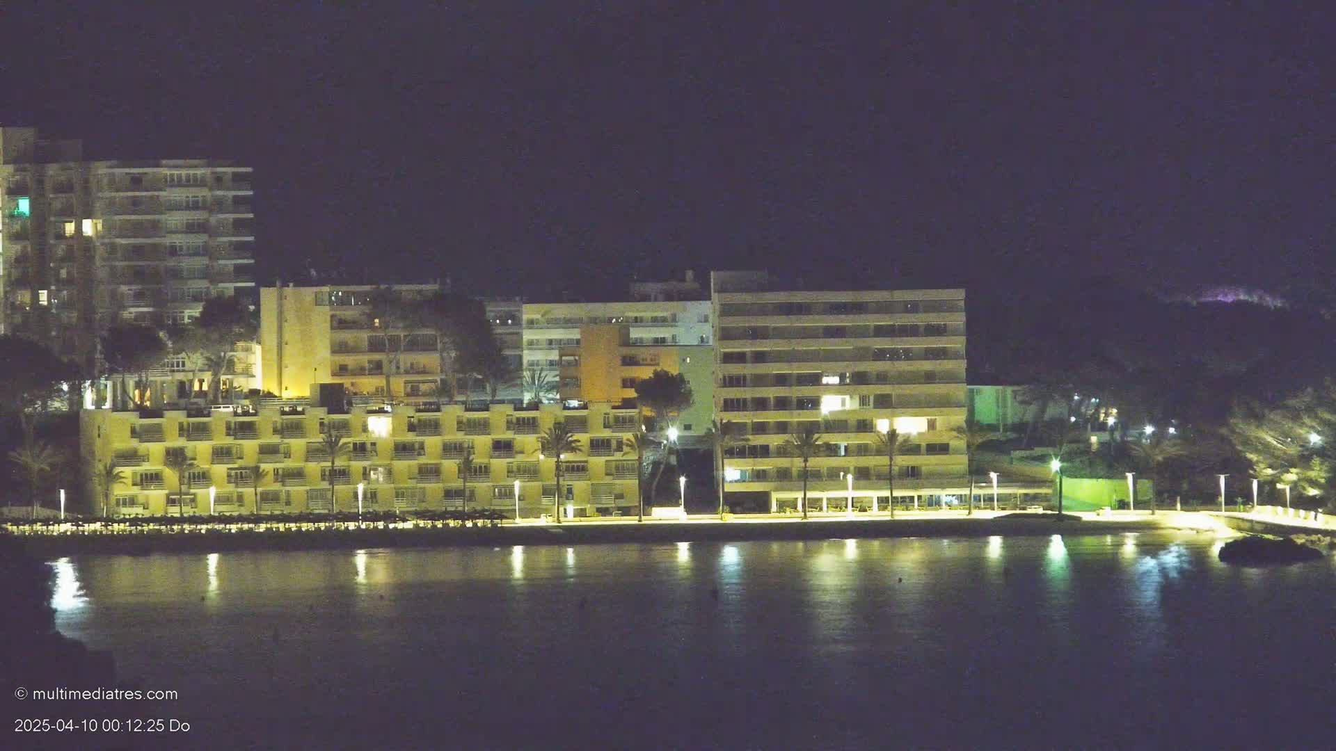 Lit-up buildings along a waterfront at night, reflecting in calm water under a dark sky.