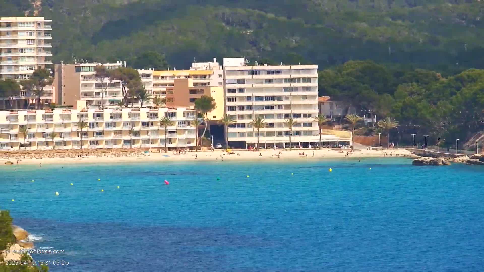A sandy beach with several multi-story buildings behind it sits on a clear, turquoise body of water, backed by a lush green hillside under a sunny sky.