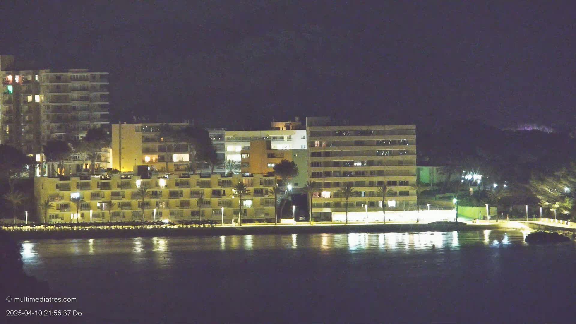 A nighttime view of illuminated multi-story buildings along a waterfront, reflected in calm water under a dark sky.