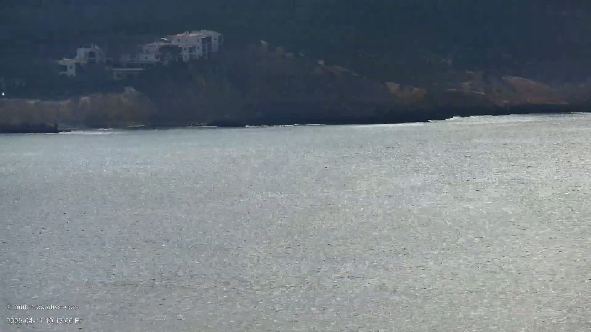 A body of water with small waves reflects sunlight, while a dark cliff with several light-colored buildings sits in the background.