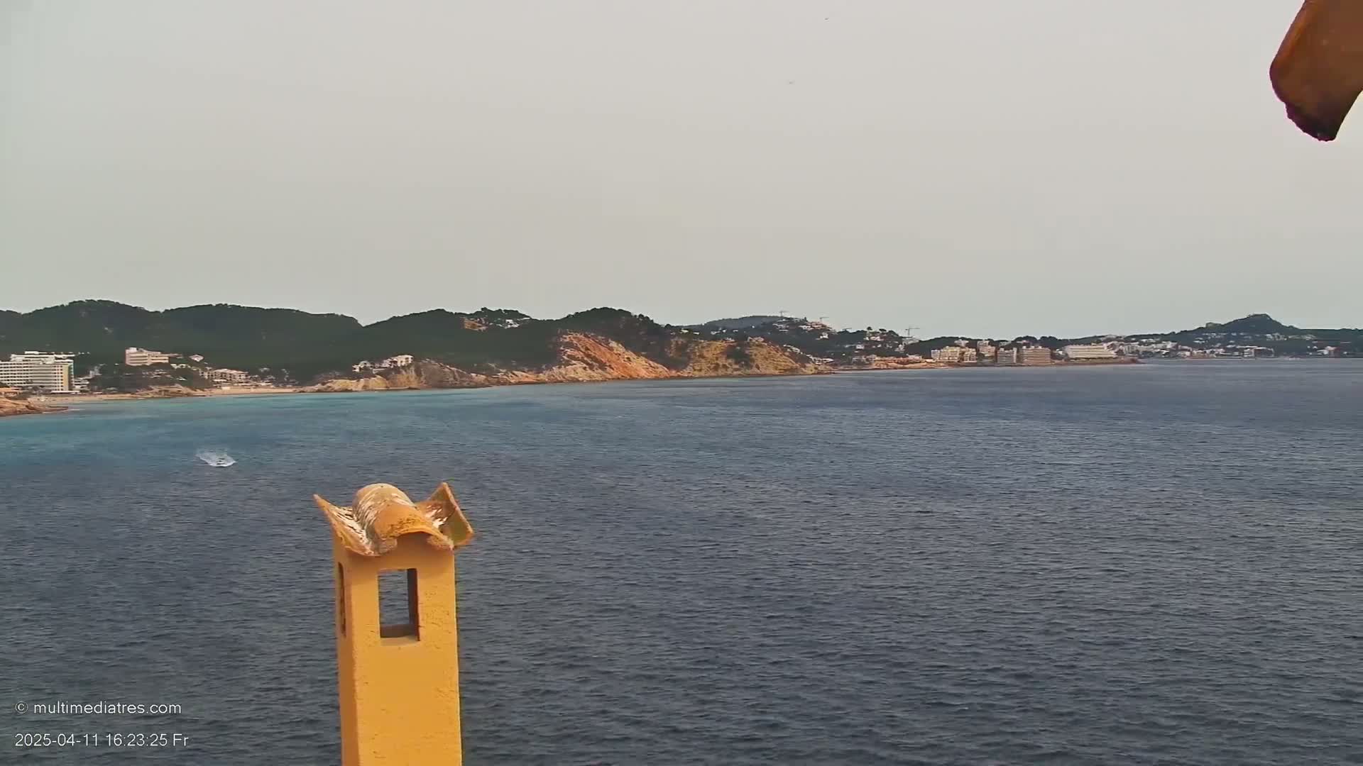 A calm sea stretches out before a hilly coastline with buildings and vegetation, viewed from behind a yellow chimney on a mostly cloudy day.