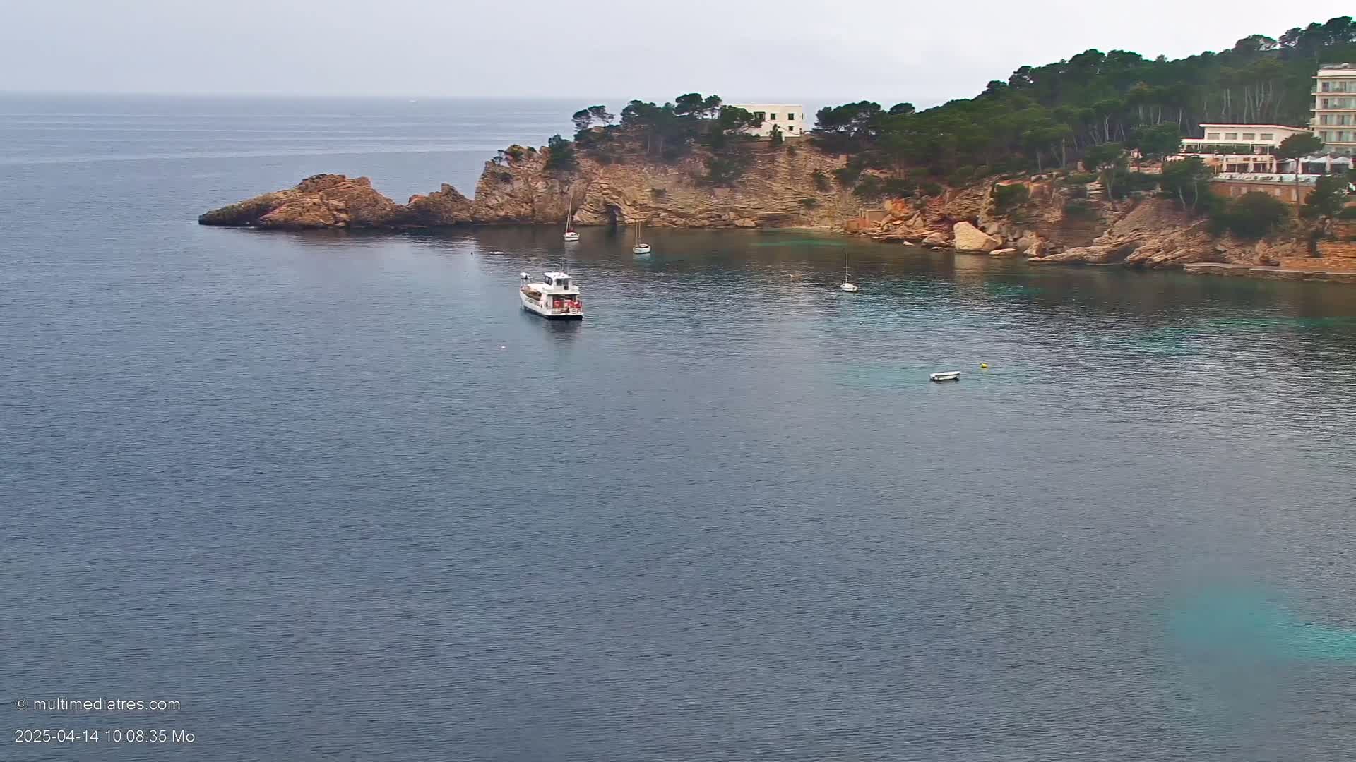 A calm, overcast day reveals a secluded cove with a few boats, a rocky shoreline dotted with vegetation and buildings, and a deep blue sea extending to the horizon.