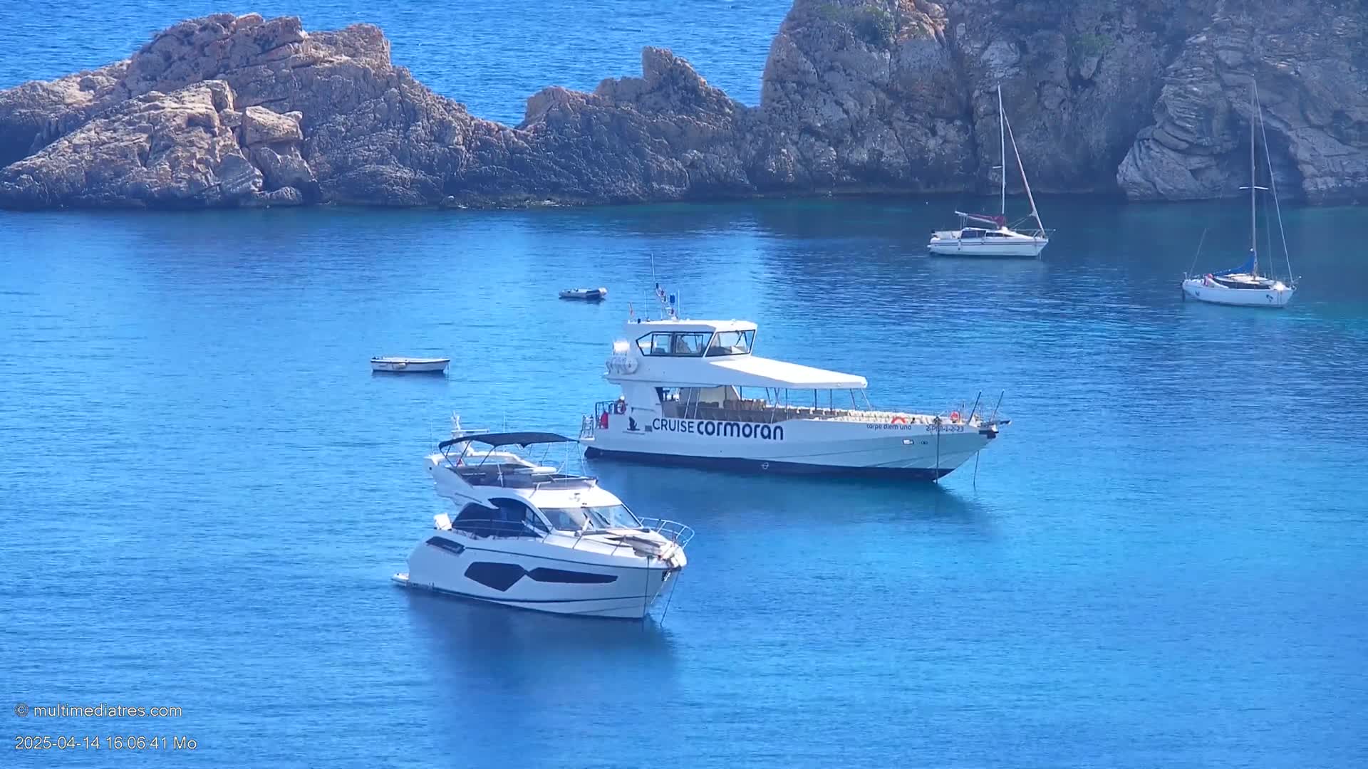 Several boats, including a large passenger vessel and a smaller motor yacht, are moored in a calm, clear blue bay surrounded by rocky cliffs under a sunny sky.