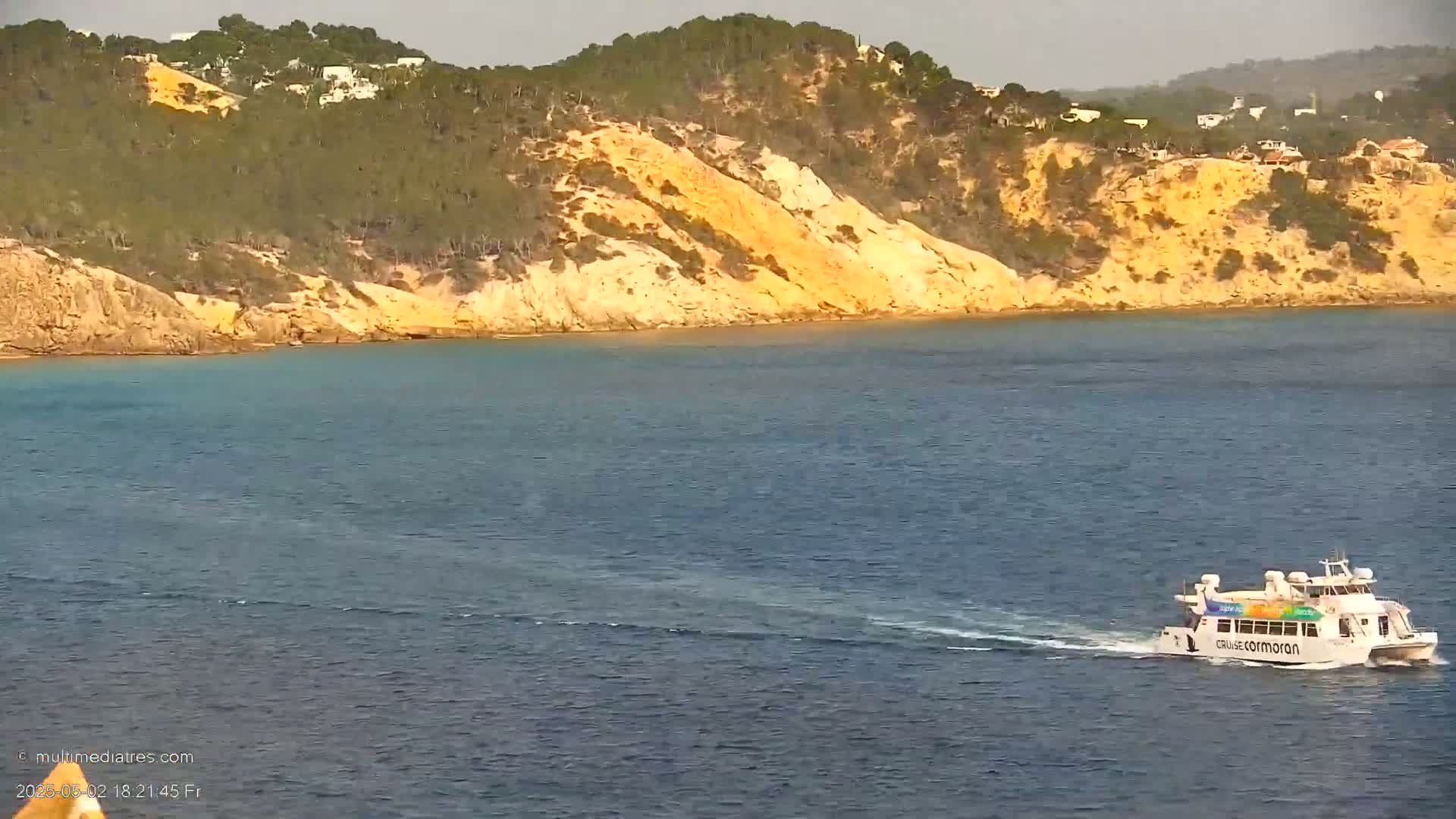 A ferry travels across calm, blue water toward a tan, tree-covered hillside.