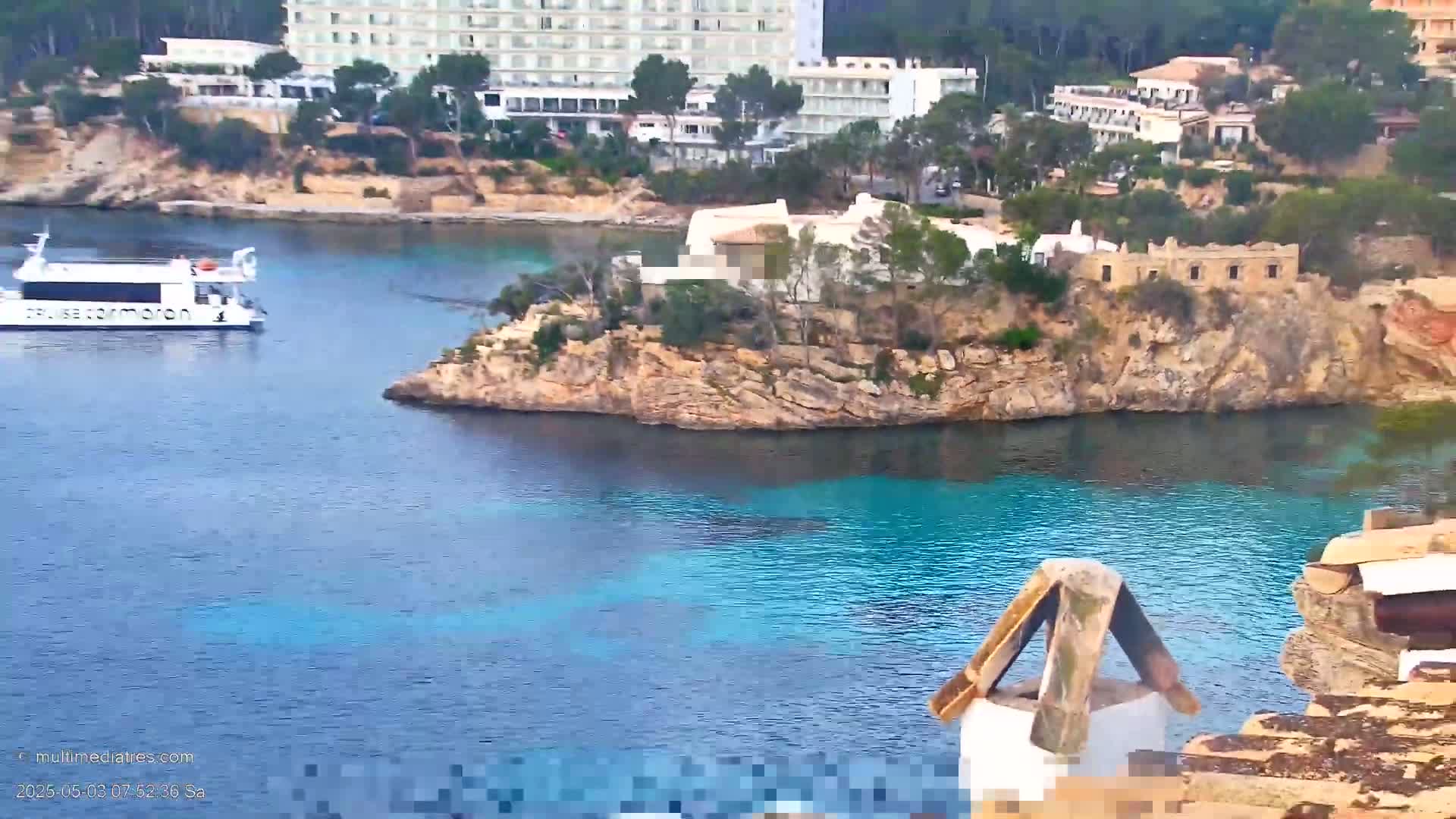 A white boat is in a calm, turquoise bay near a rocky shoreline with buildings and trees on the hillside.