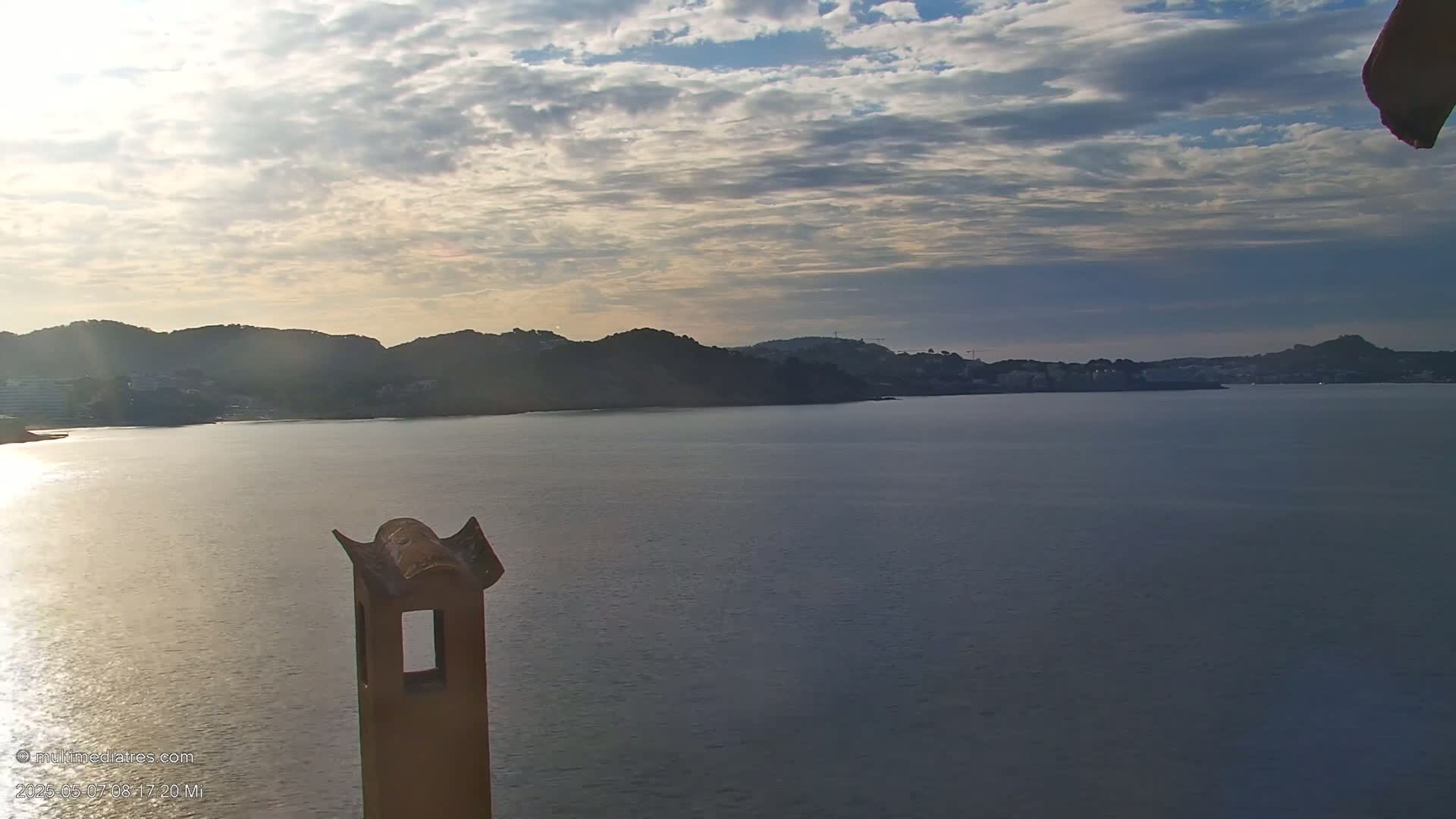 A calm body of water stretches towards a distant shoreline of dark hills under a partly cloudy sky.
