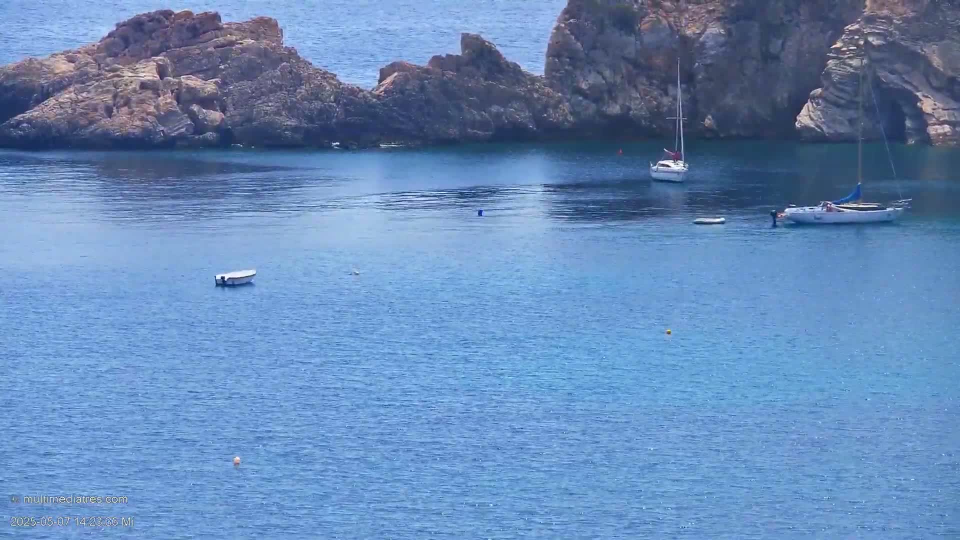 Calm blue water laps against rocky shores where several small boats are anchored.