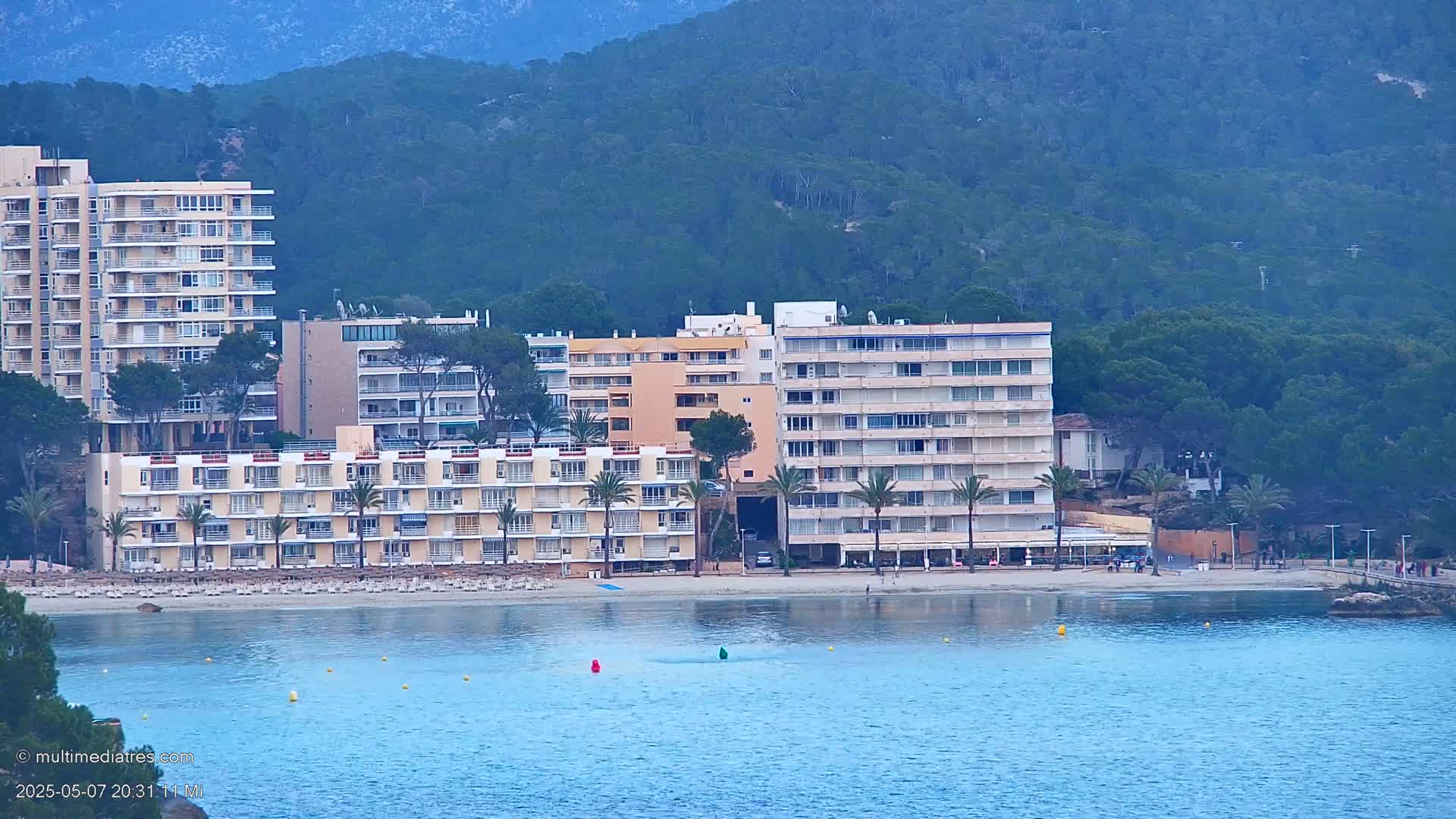 Several multi-story hotels line a sandy beach next to calm blue water and a background of a dark green mountain.