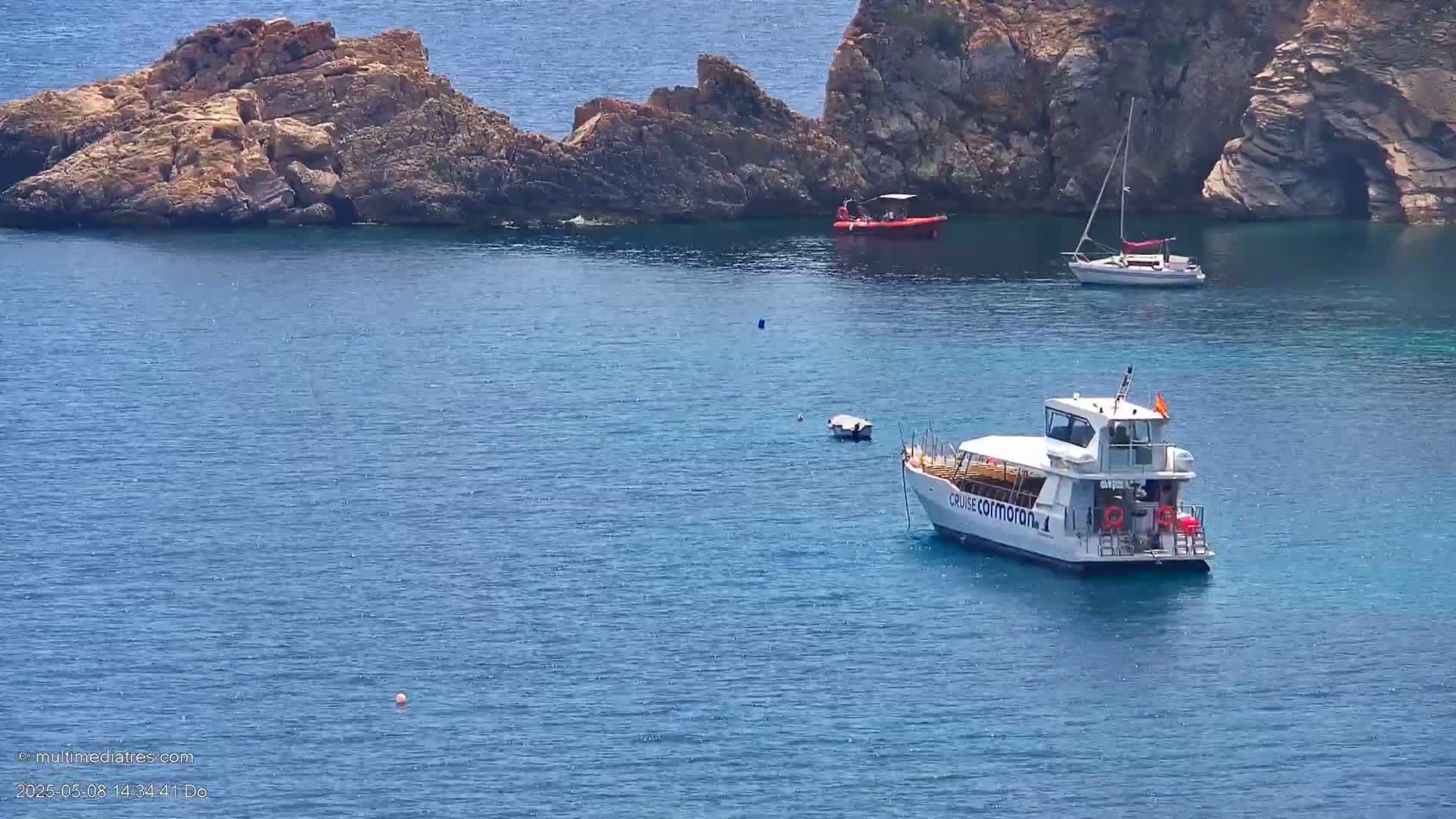 A calm blue sea surrounds rocky outcrops, with a passenger boat, a small sailboat, and a rigid-hulled inflatable boat visible.