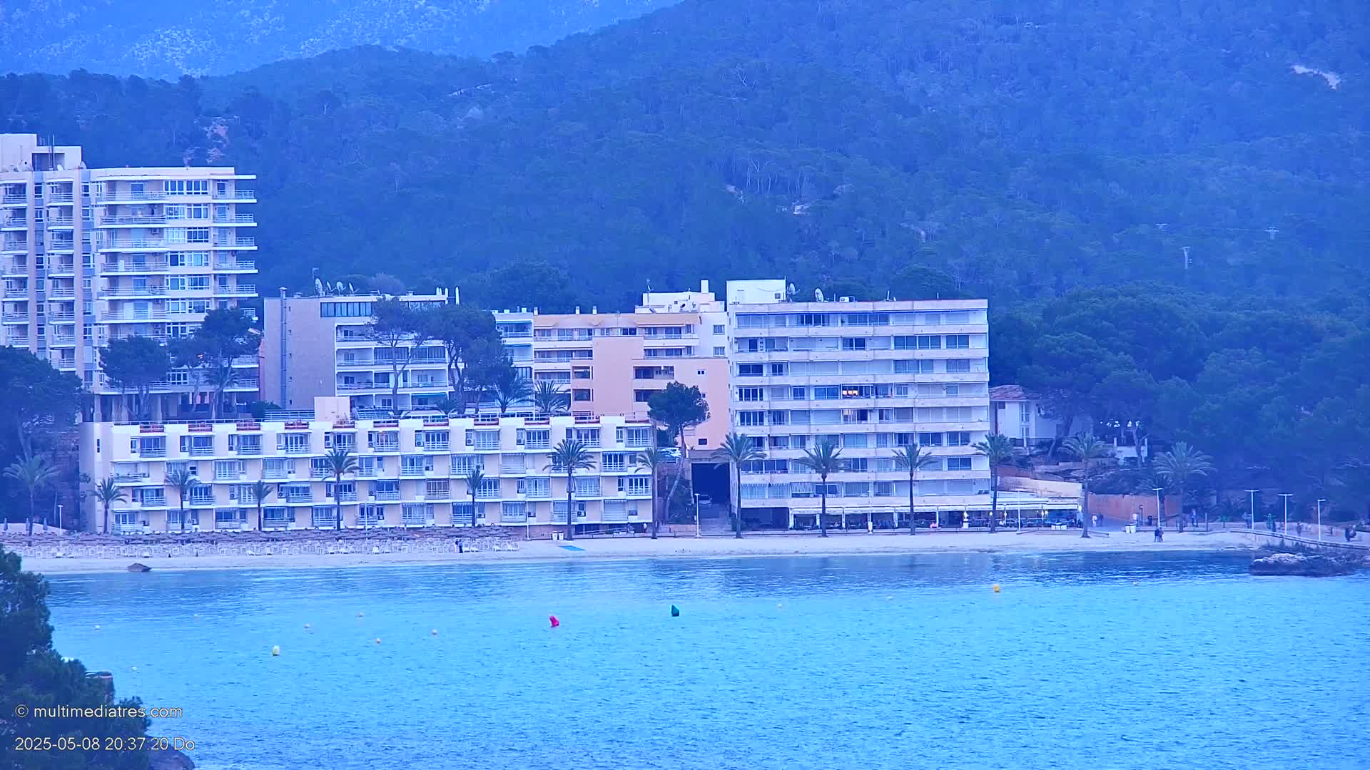 Several multi-story hotels line a beach next to calm blue water, backed by a hazy blue mountain range.