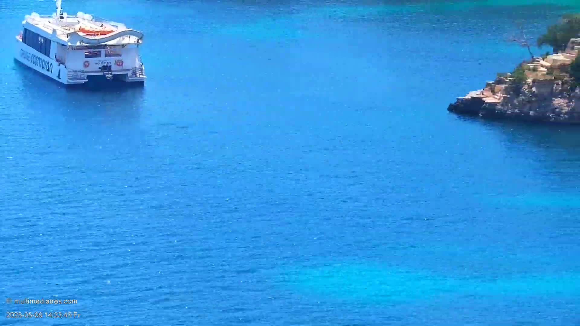 A white catamaran floats on calm, bright blue water near a rocky shoreline.
