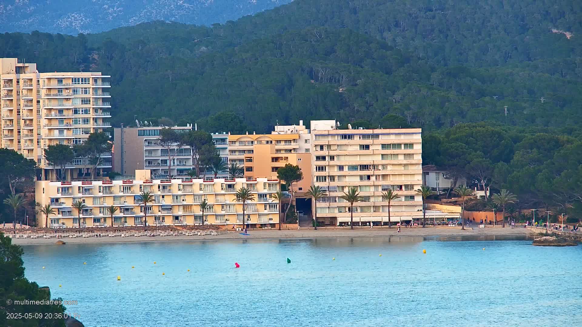 Several multi-story hotels sit on a sandy beach next to calm, blue water, backed by a lush, green hillside.