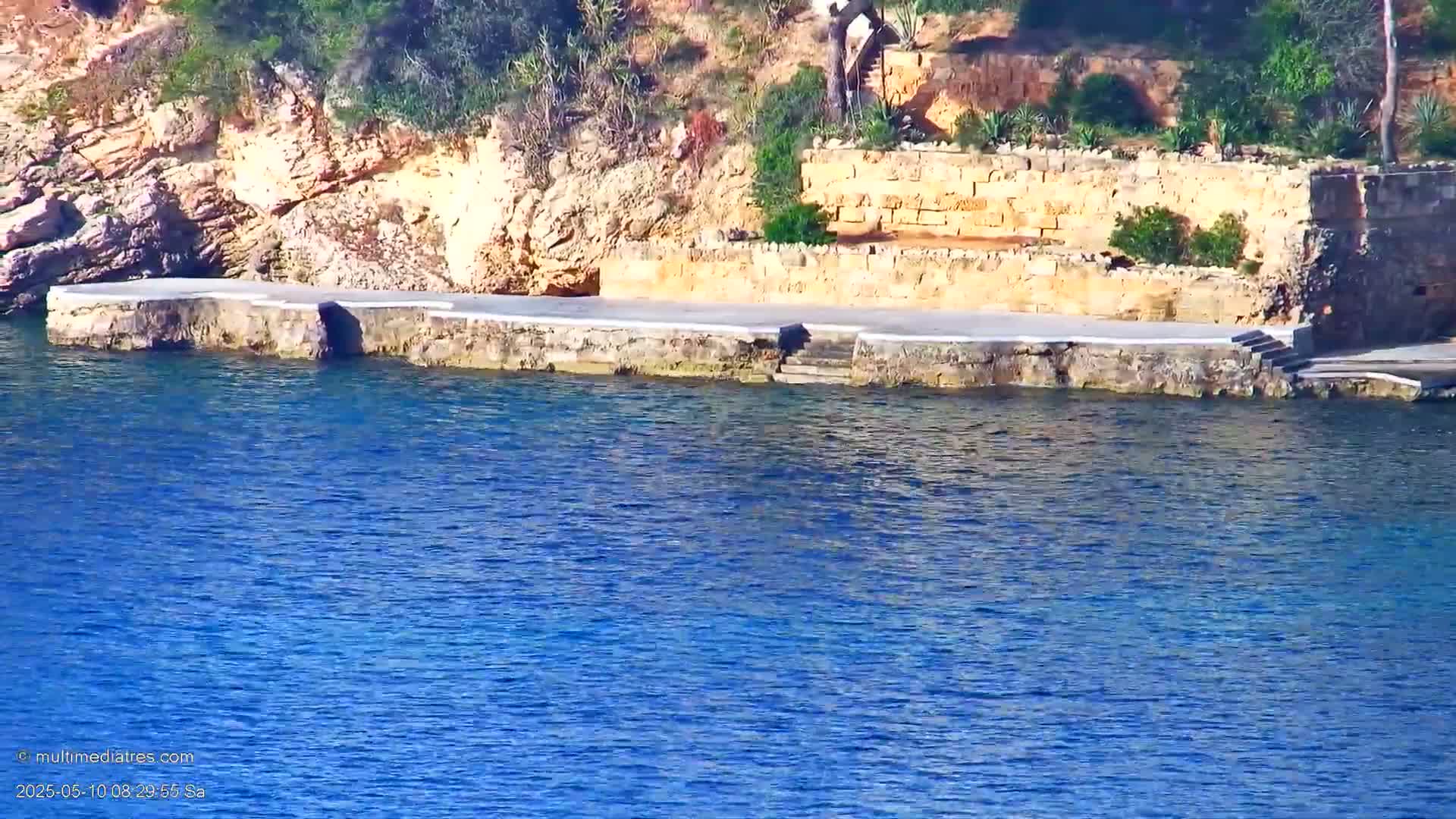 A stone platform with steps leading into clear blue water sits against a rocky hillside with sparse vegetation under a sunny sky.