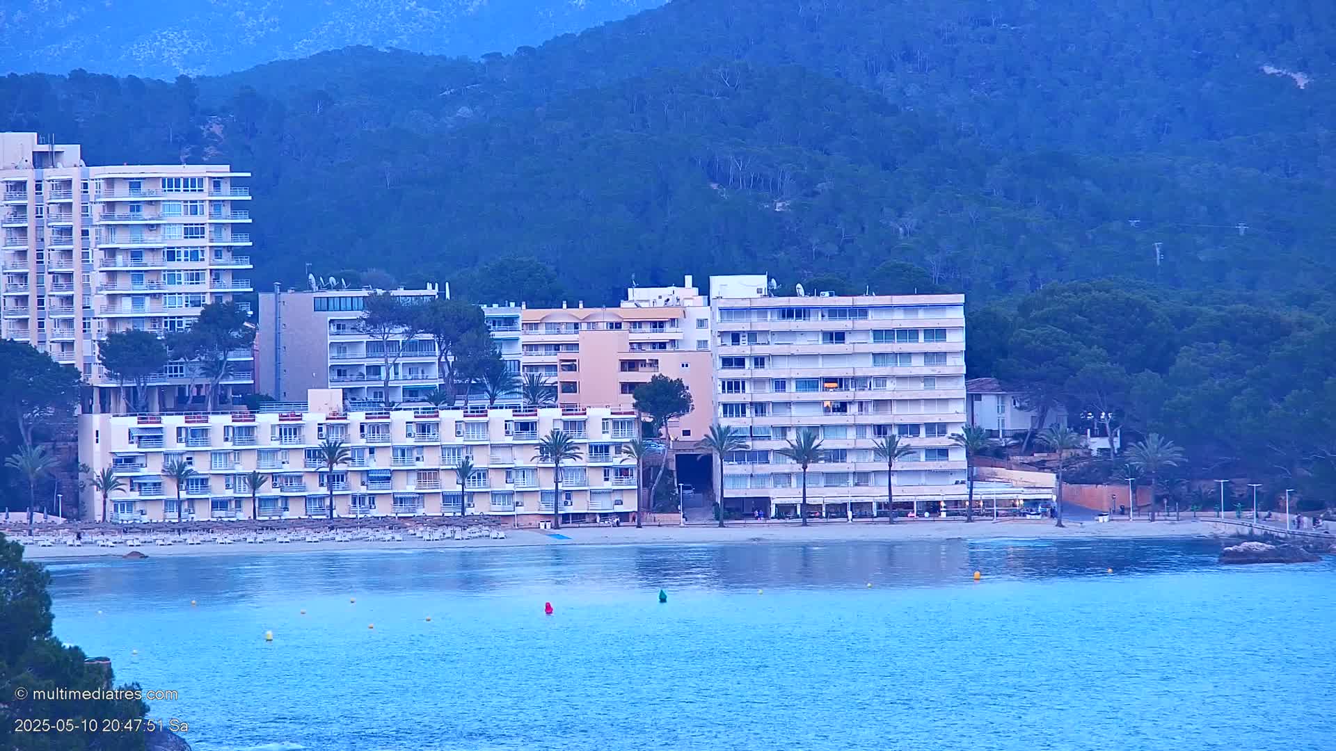 Several multi-story buildings line a beach with calm blue water, backed by a dark blue, forested hillside.