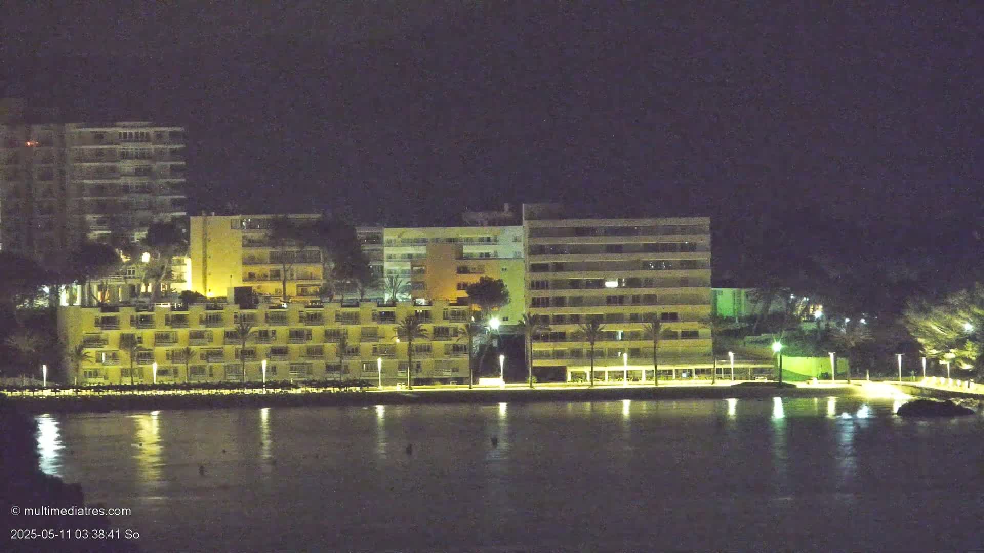 Illuminated multi-story buildings line a calm body of water at night.