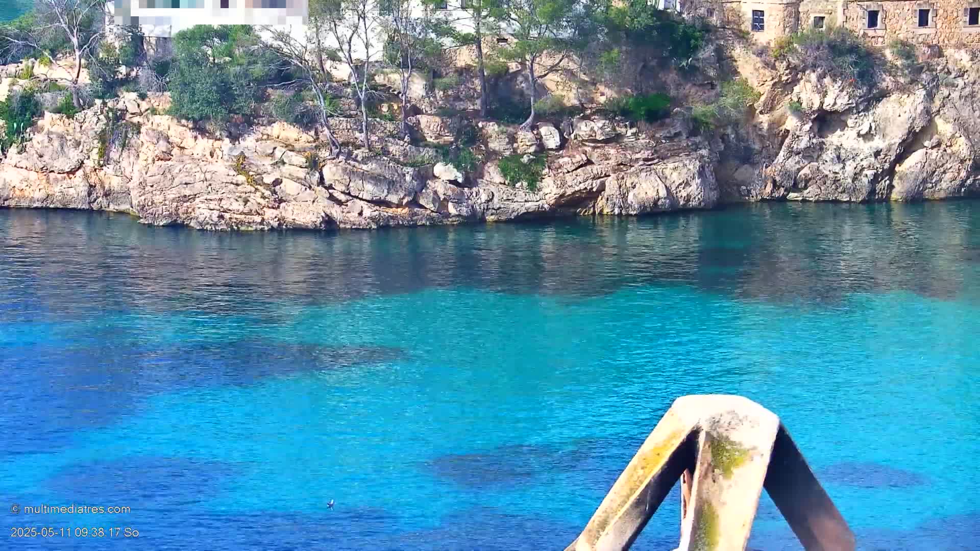 Brightly turquoise water laps against a rocky shore where a few trees grow atop a stone cliff near a building.