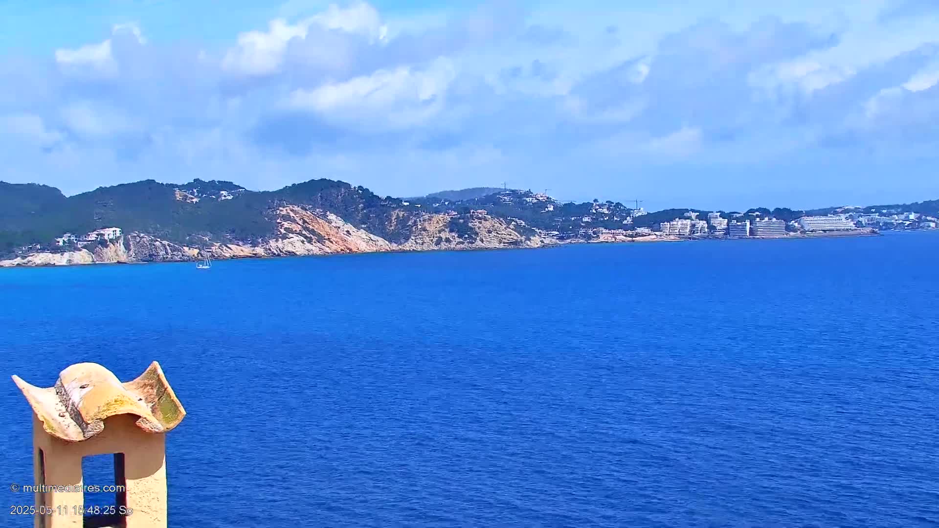 A sailboat is visible on a calm, blue sea in front of a rocky, green coastline with buildings in the distance under a partly cloudy sky.