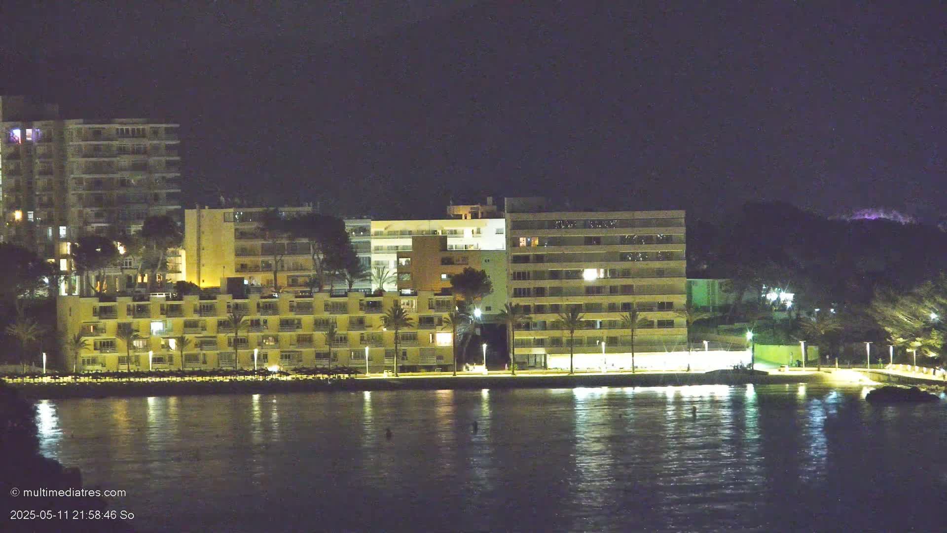 Illuminated buildings line a waterfront at night, reflected in calm water under a dark sky.
