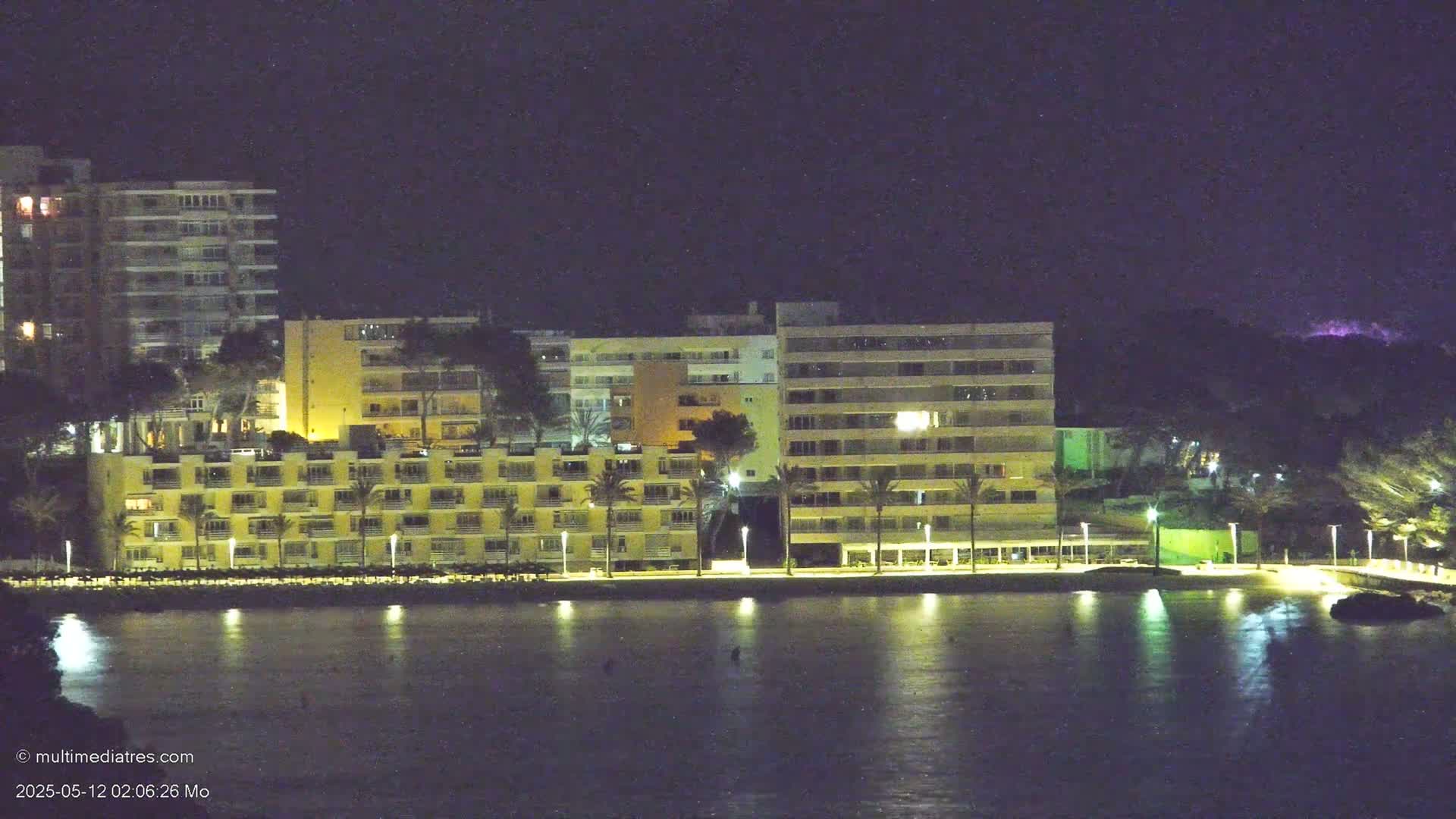 Lit-up apartment buildings line a calm, dark waterfront at night.