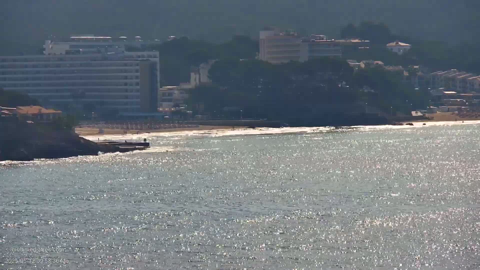 Sparkling water laps a sandy beach with buildings and hotels situated on a hillside in the background on a sunny day.