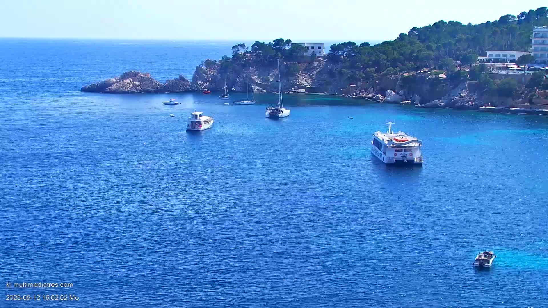Several boats are anchored in a calm, blue ocean cove next to a rocky shore lined with green trees and buildings.