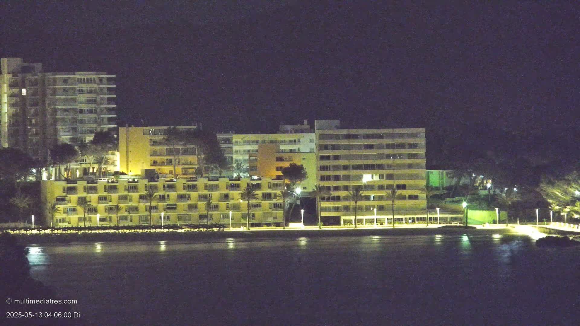 At night, several illuminated apartment buildings line a waterfront, palm trees dot the foreground, and calm water reflects the lights.