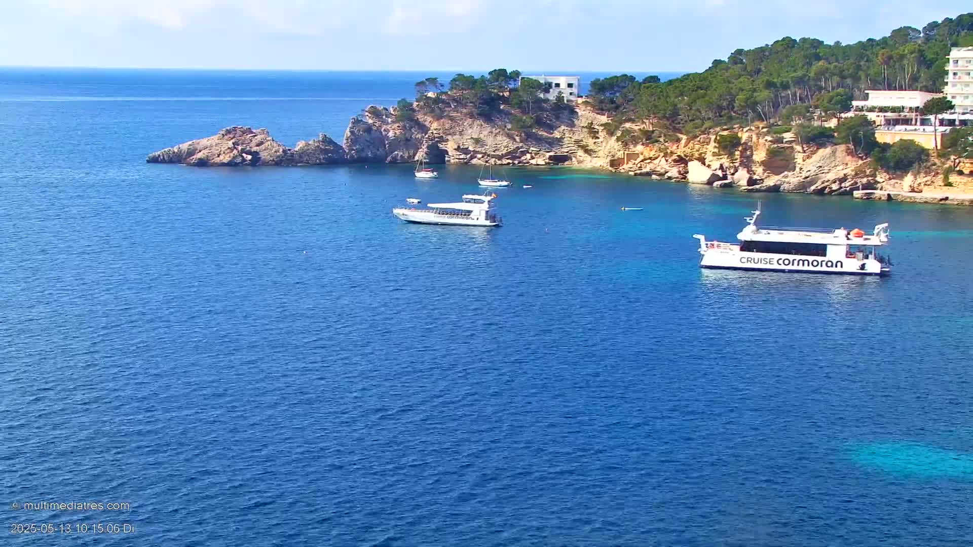 Several boats are anchored in a calm, blue sea near a rocky, tree-covered coastline on a sunny day.