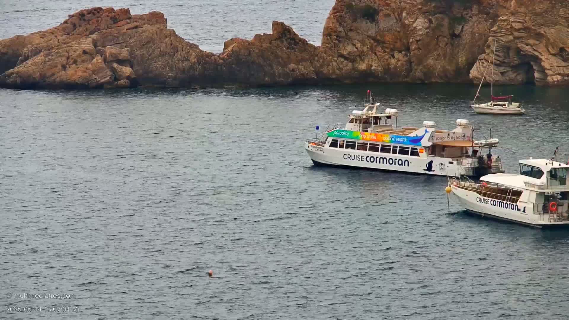 Two tour boats are moored near rocky cliffs in calm water; a small sailboat is further away.
