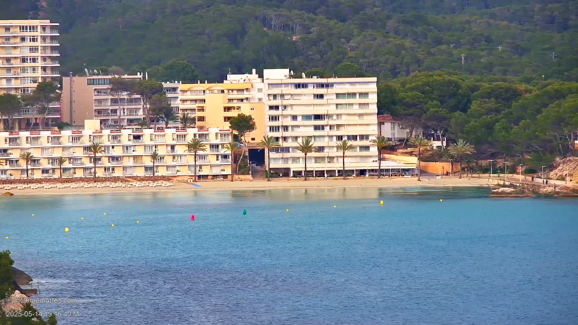 A beach with several multi-story hotels sits on a calm, clear body of water bordered by lush green hills.
