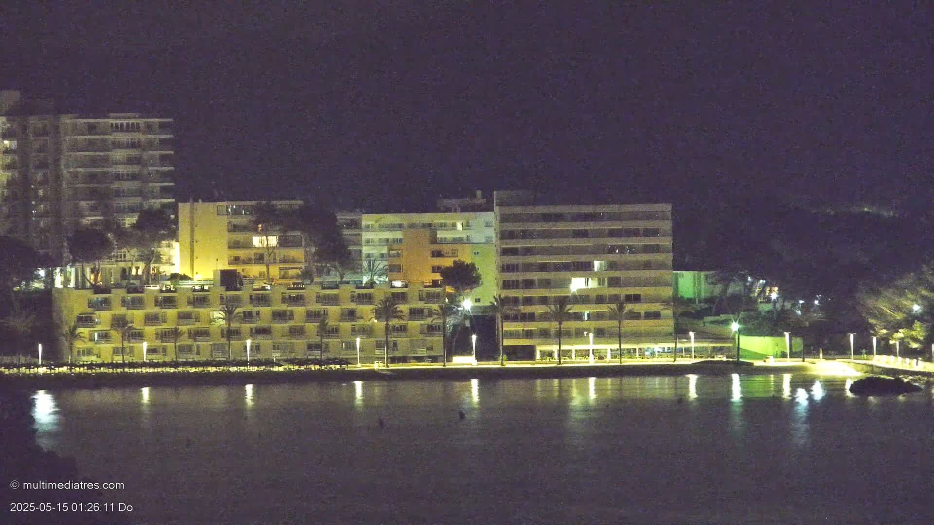 Illuminated multi-story buildings line a calm body of water at night.