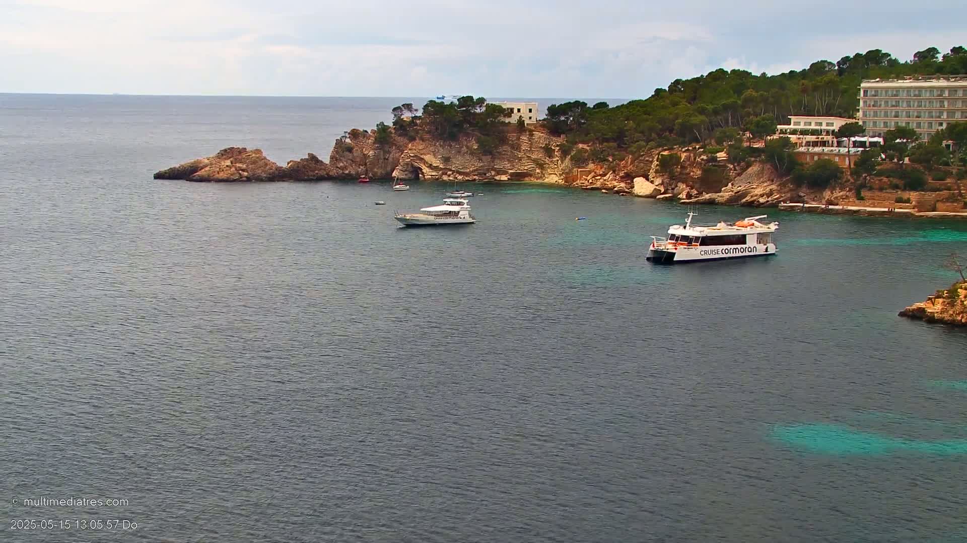A calm sea with several boats near a rocky coastline featuring a hotel and lush green vegetation under an overcast sky.
