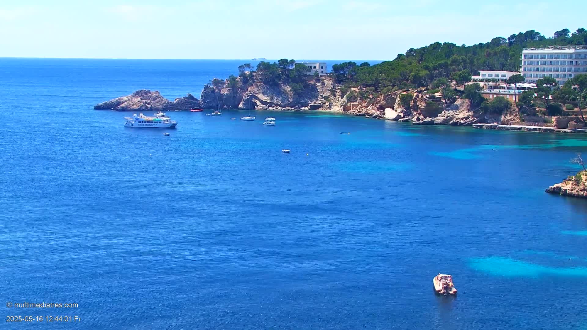 A calm, sunny day shows a tranquil blue ocean cove with several boats, a small passenger ferry, rocky cliffs covered in green trees, and a large white hotel.
