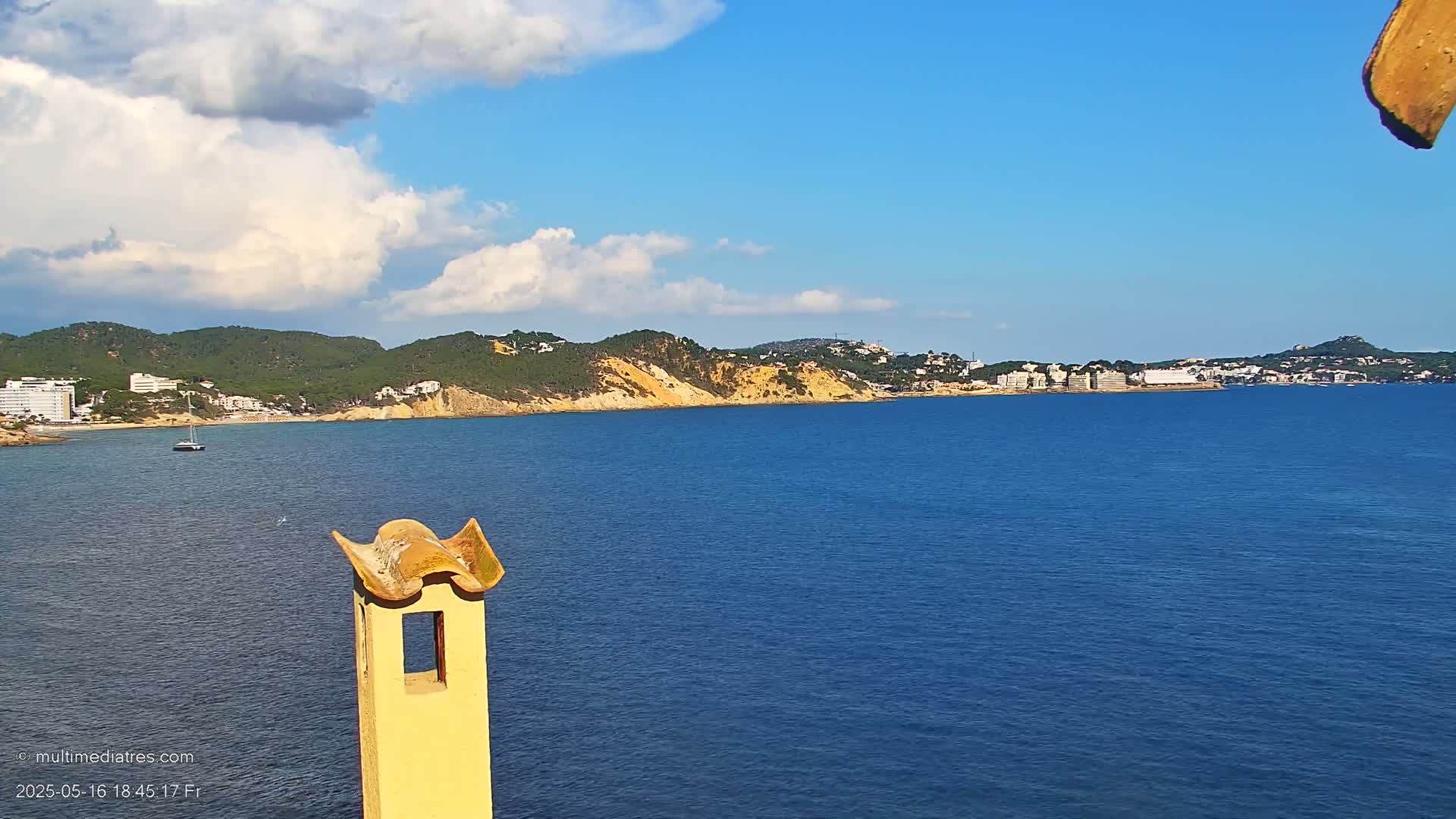 A partly cloudy blue sky overlooks a calm blue sea with a distant coastline of green hills and buildings, partially obscured by a yellowish chimney in the foreground.