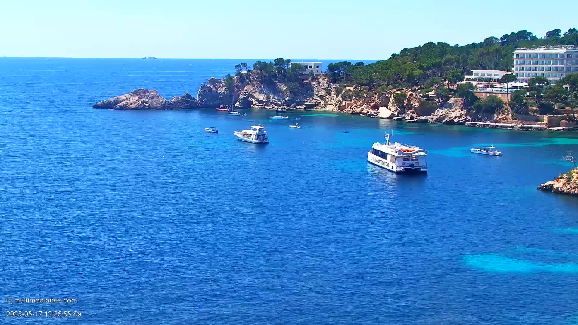 A calm, sunny day shows several boats in a clear blue bay surrounded by rocky shores and a hotel.