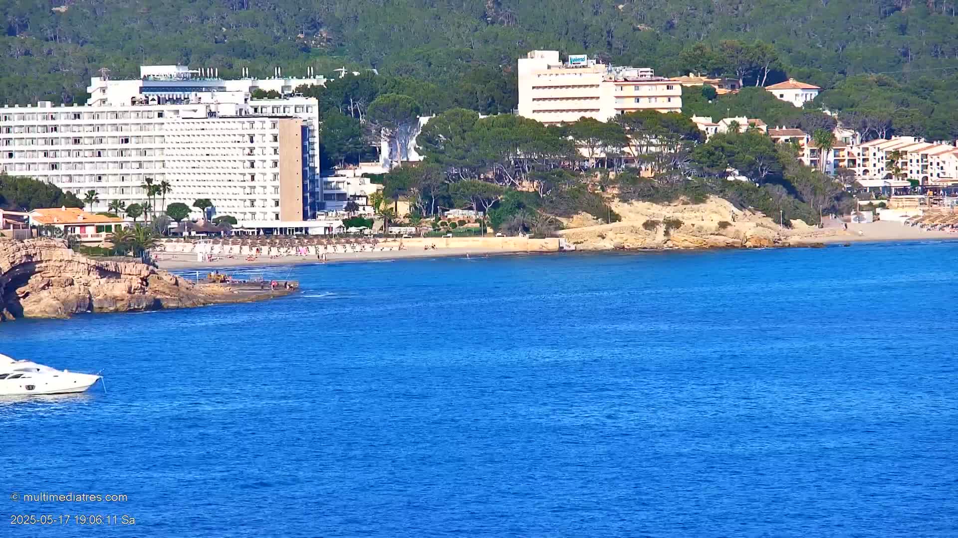 A sandy beach with several hotels and buildings sits on a coastline with blue water and a white boat in the foreground on a sunny day.