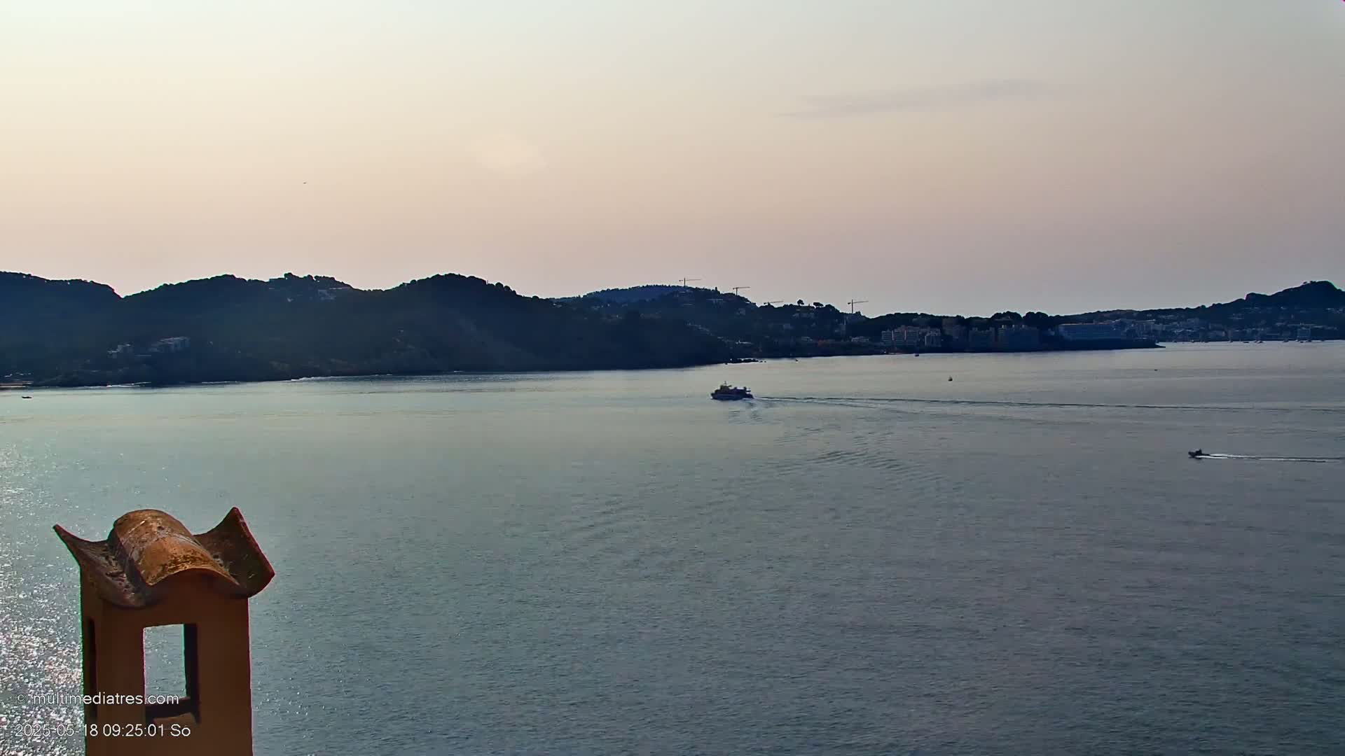 A calm body of water with a few small boats,  viewed from a high vantage point overlooking a distant shoreline of hills and buildings under a pale, dusky sky.