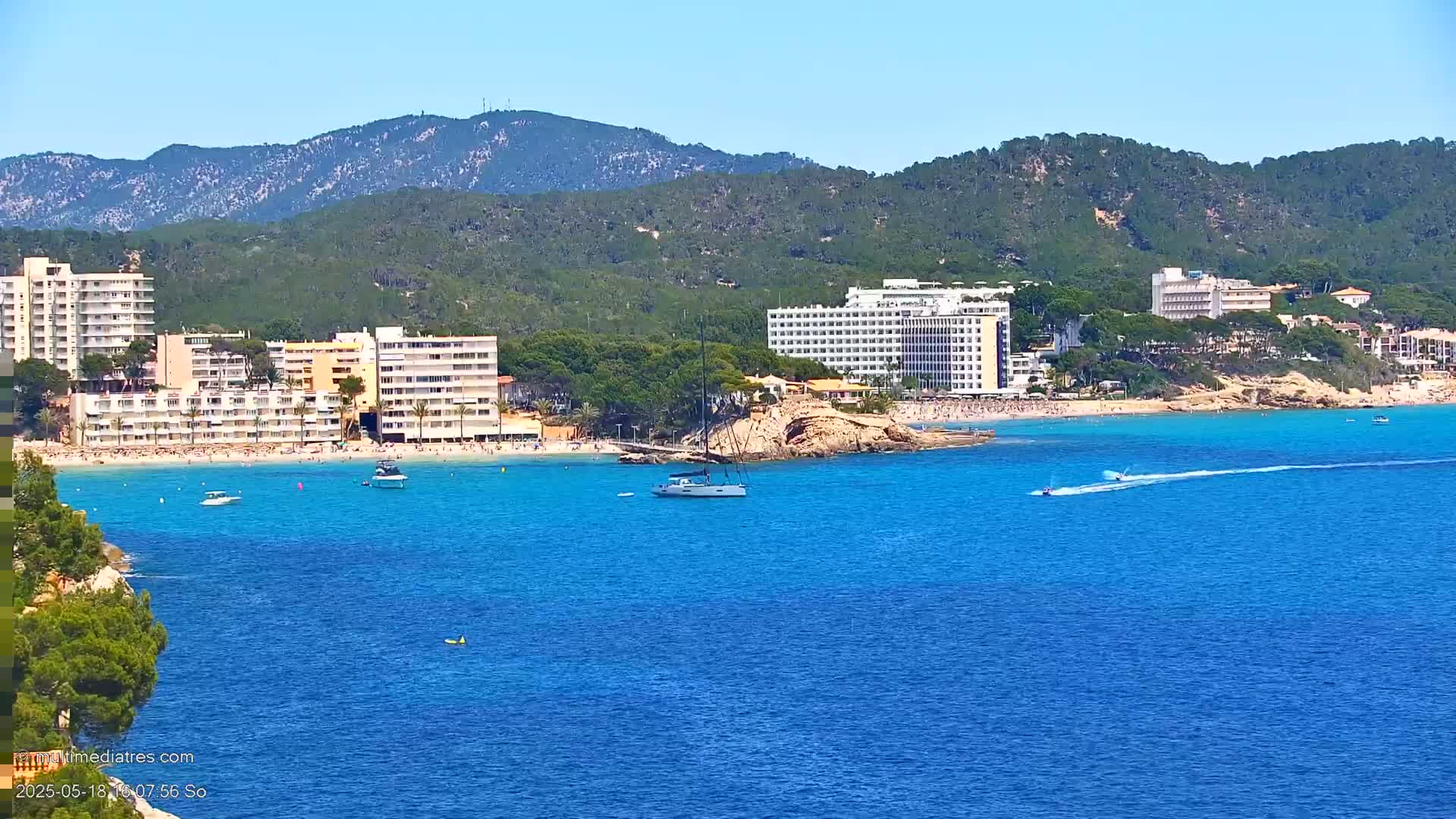 A sunny day at a beach with turquoise water shows several large hotels and many people relaxing on the shore, with boats and jet skis in the water, and mountains in the background.