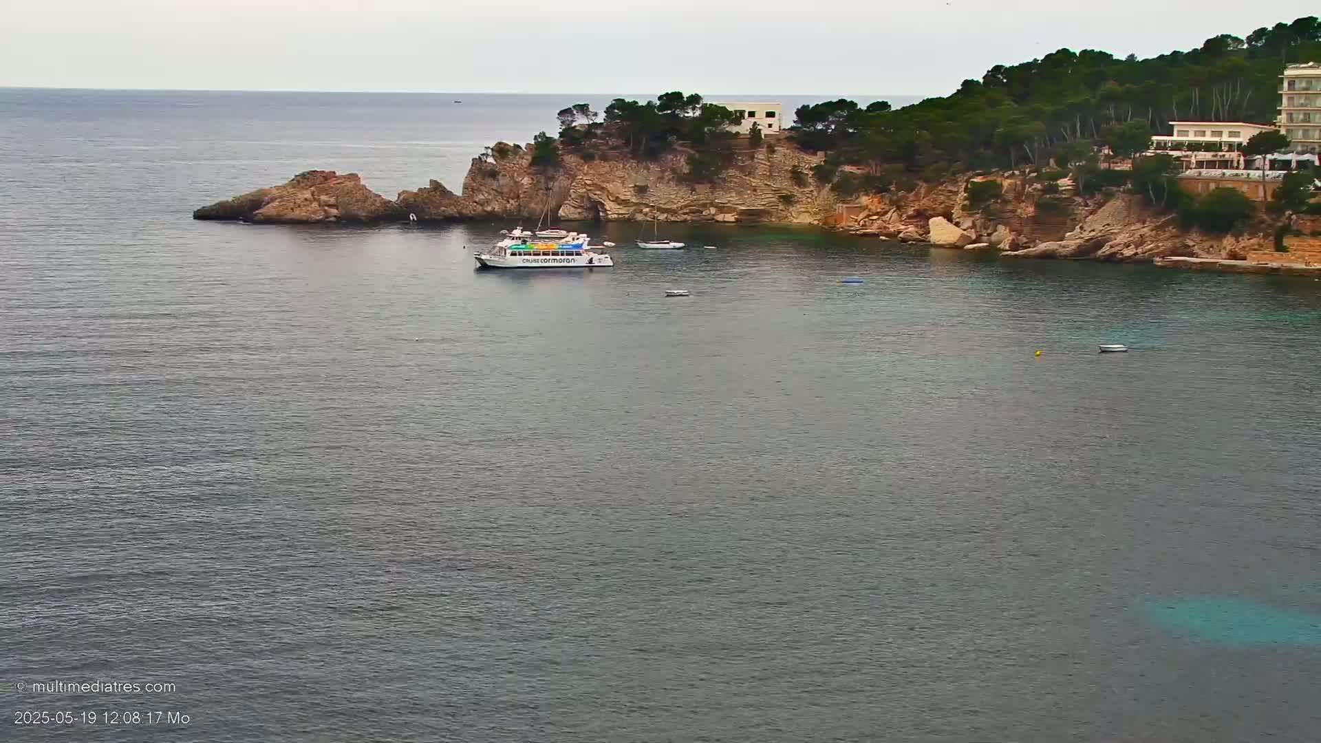 A boat is anchored in a calm bay next to a rocky coastline with green trees and buildings on top.
