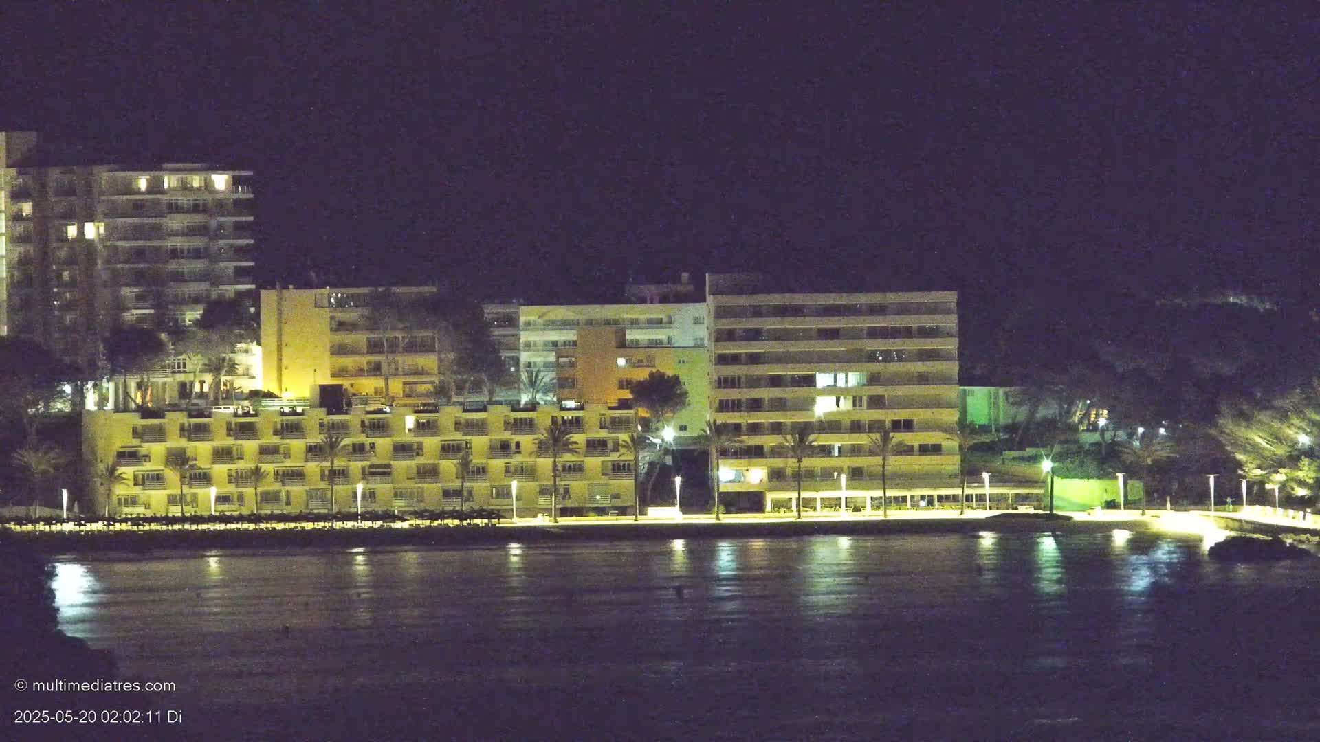 A nighttime view of several illuminated beachfront hotels and palm trees, reflected in the calm water.
