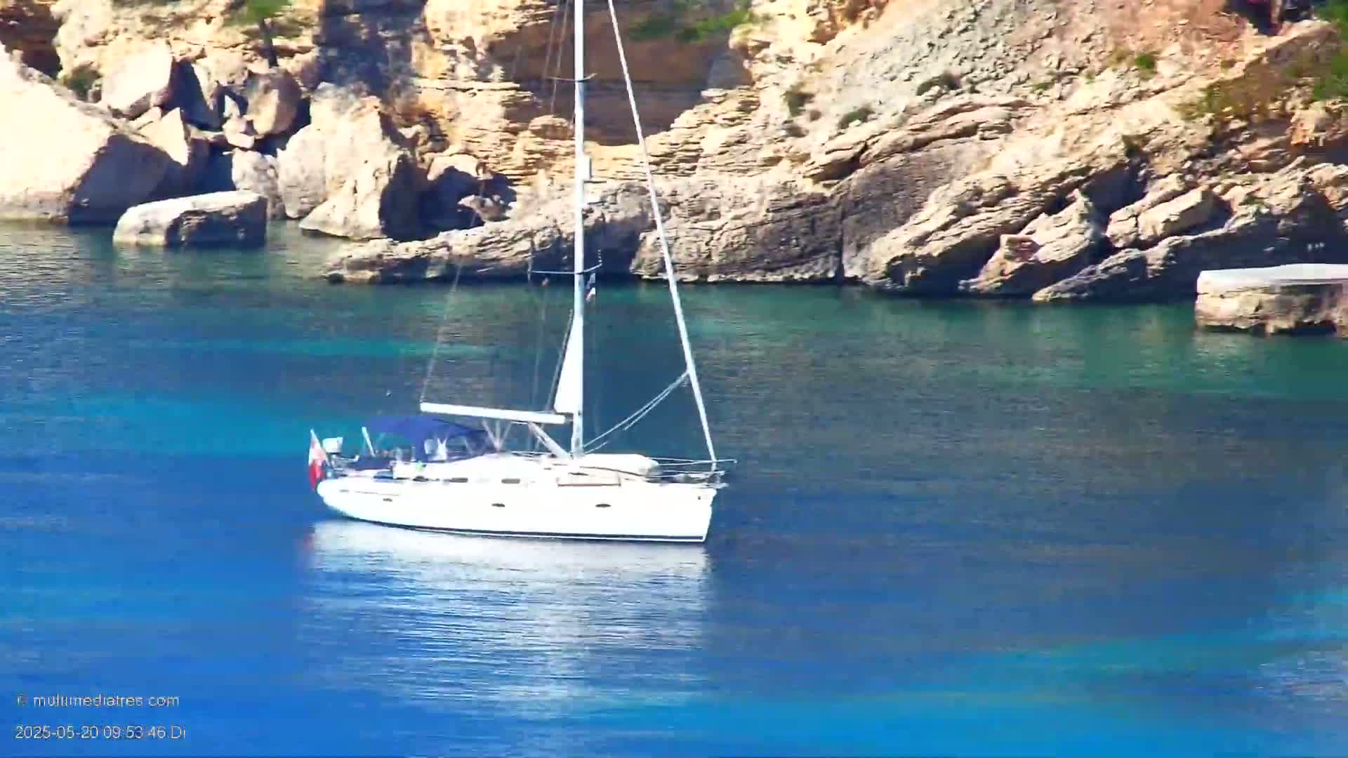 A white sailboat is anchored in calm, clear blue water near rocky cliffs on a sunny day.