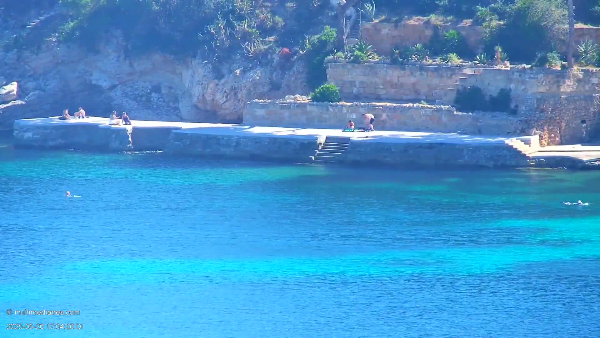 Several people relax and swim in clear, turquoise water near a stone platform and steps that lead down from a rocky, sunlit shoreline.
