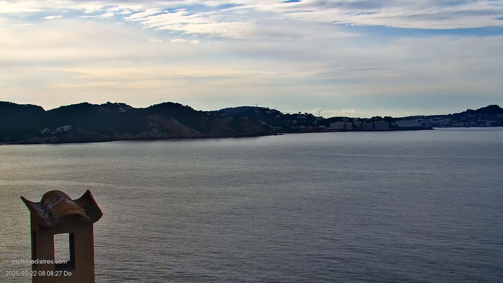 A calm body of water stretches out to a distant coastline under a partly cloudy sky, with a portion of a stone chimney visible in the foreground.