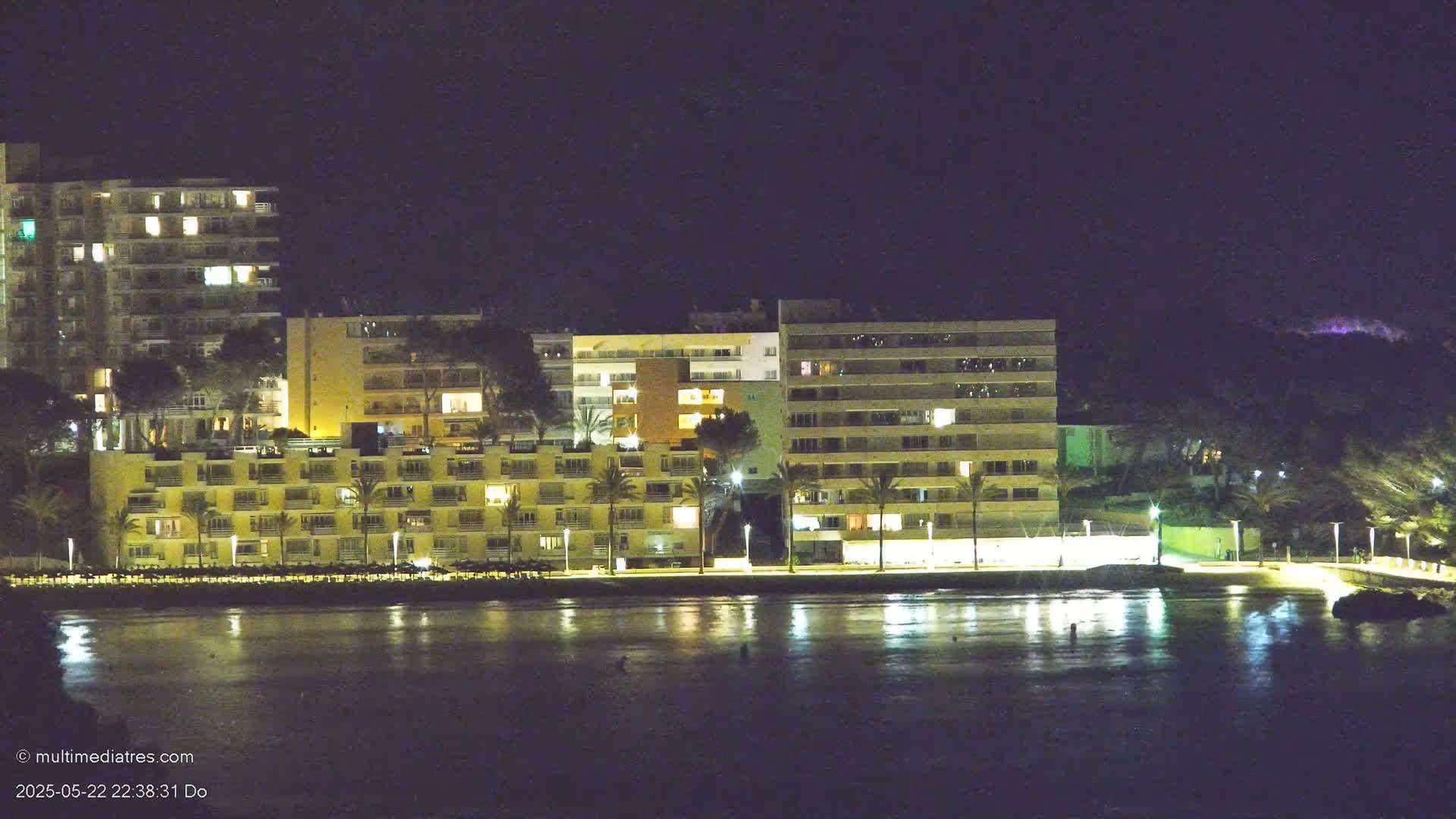 A row of illuminated apartment buildings sits on a calm, dark body of water at night.