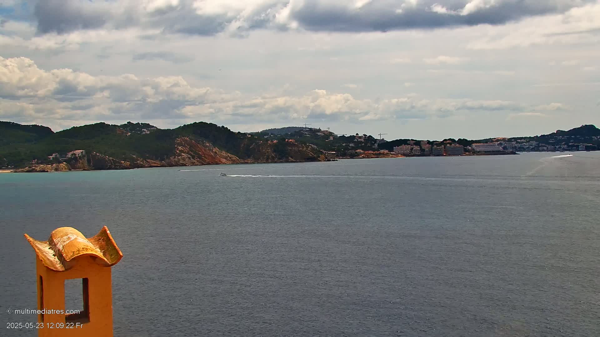 A partly cloudy day overlooks a calm body of water with a boat, a hilly coastline with buildings, and a portion of a yellow chimney in the foreground.
