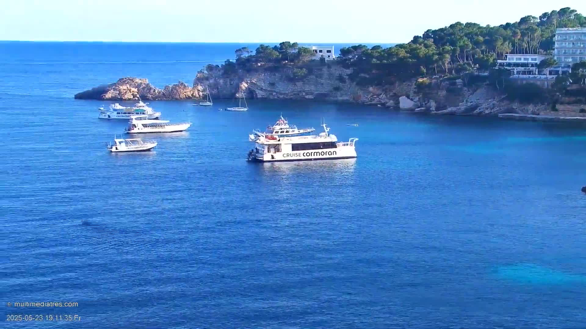 Several boats are anchored in a calm, blue ocean cove near a rocky shore lined with green trees and buildings.