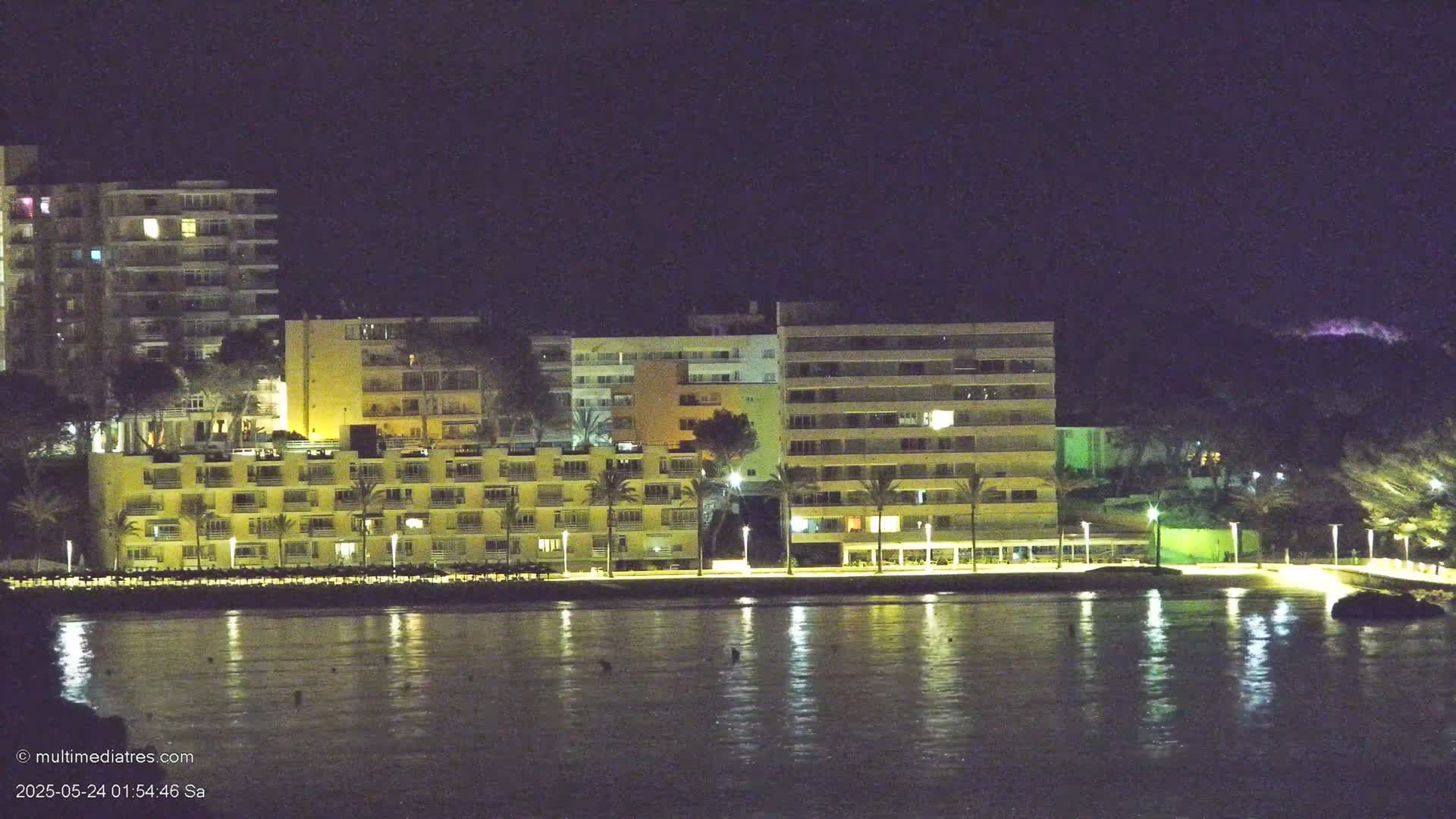 Illuminated beachfront buildings and palm trees are reflected in the calm nighttime waters.