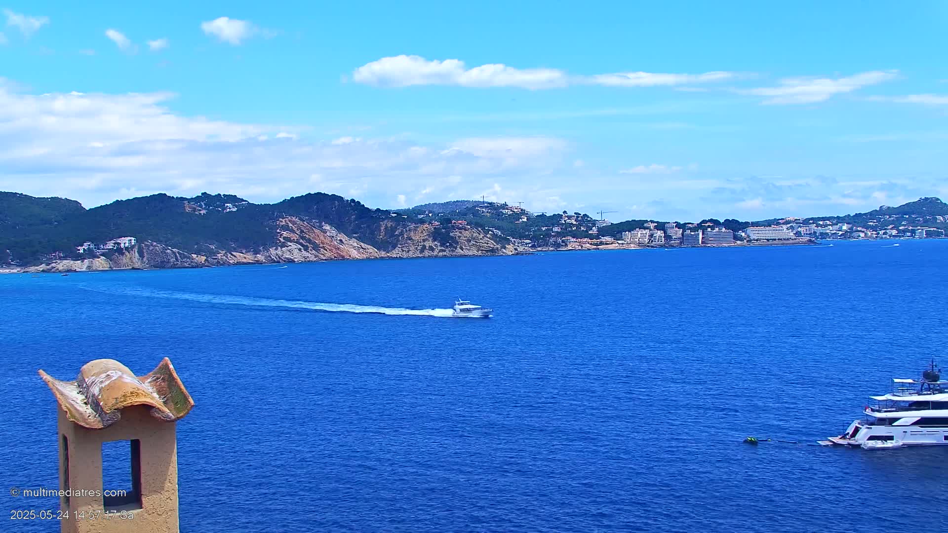 A blue sea with two boats, one small and one large, under a mostly sunny sky with some clouds, with a green and brown hilly coastline in the background.