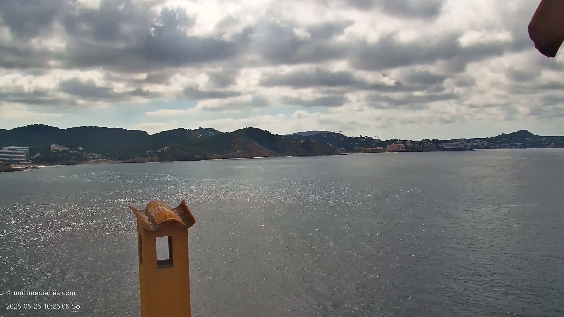 A partly cloudy day reveals a coastal view of a calm sea with a distant shoreline featuring buildings and hills, partially obscured by a chimney in the foreground.