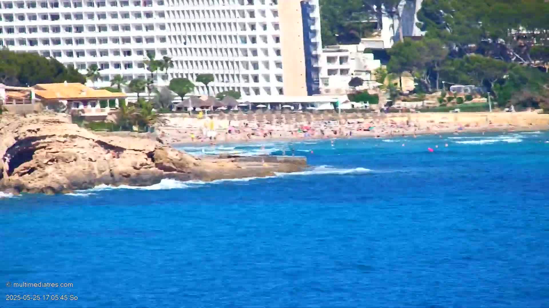 A sunny day at a beach with a large white hotel in the background, rocky outcrops in the foreground, and many people enjoying the sand and water.